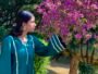 Woman wearing a teal green kurta with rainbow silver lace detailing and flared sleeves beside pink flowers in a garden