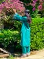 Back view of a woman wearing a teal green kurta set with rainbow silver lace sleeve detailing beside pink flowers and green foliage