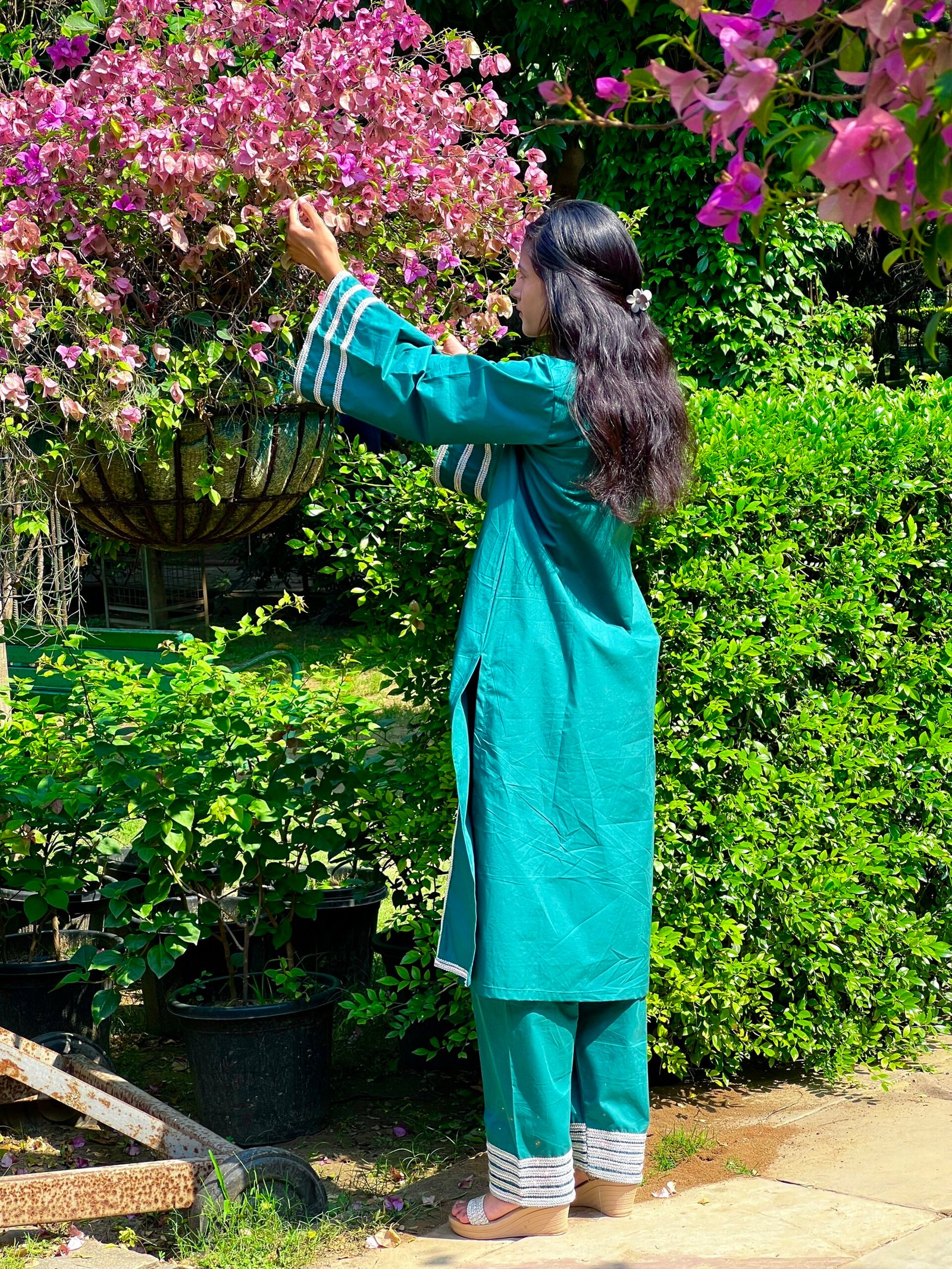 Teal Green Kurta Set – Back View with Flared Sleeves Back view of a woman wearing a teal green kurta set with rainbow silver lace sleeve detailing beside pink flowers and green foliage